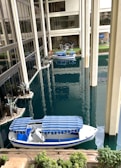 A waterway runs through the interior of a modern building, featuring a small white and blue boat with a striped canopy. The building is characterized by large glass windows and tall columns, with landscaped areas including small plants and decorative stones. In the background, there are outdoor seating arrangements with tables and chairs.