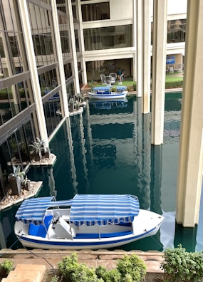 A waterway runs through the interior of a modern building, featuring a small white and blue boat with a striped canopy. The building is characterized by large glass windows and tall columns, with landscaped areas including small plants and decorative stones. In the background, there are outdoor seating arrangements with tables and chairs.