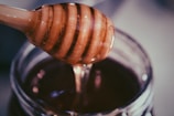 A wooden honey dipper coated with golden honey rests above a jar filled with rich, dark honey. The light catches the viscous texture of the honey, highlighting its glistening surface.