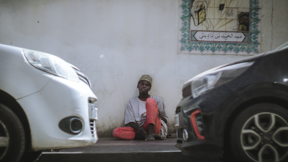 a man sitting on the side of a building next to parked cars
