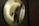 A close-up of a brass-colored door latch with intricate textured detailing. The latch is set in a dark brown door, and the lighting highlights the metallic surface.