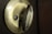 A close-up of a brass-colored door latch with intricate textured detailing. The latch is set in a dark brown door, and the lighting highlights the metallic surface.