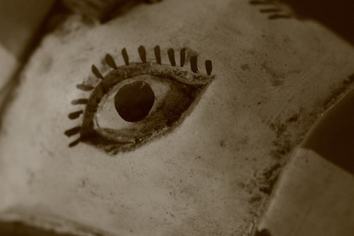 A close-up view of a carved eye on a sculpture or mask, with prominent black lashes and a circular black pupil. The material appears weathered and textured, giving an aged appearance.