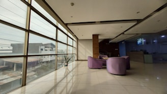 A spacious, modern interior with large windows allowing natural light to flood in, showing an urban landscape outside. The room features sleek, minimalist design elements with a polished tiled floor and a few purple chairs arranged in a semi-circle. There's a small potted plant near the window and a reception desk is visible in the background.