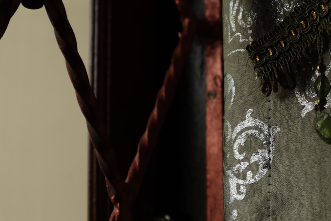 A detailed view of a decorative fabric with ornate silver patterns and fringes. A twisted rope is visible on the left side, set against a dark background.