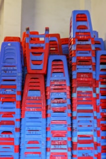 A large stack of plastic chairs arranged neatly against a wall. The chairs are predominantly blue and red, with each stack reaching considerable height, displaying a pattern created by alternating colors. The letters 'NTL' are visible on multiple chairs.