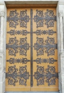 An intricately decorated wooden door featuring ornate wrought iron designs. The decorative patterns include swirling motifs and floral embellishments, creating an elaborate and detailed appearance. The door is set within a stone frame with carved columns on either side, adding to the overall grandeur.