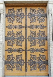 An intricately decorated wooden door featuring ornate wrought iron designs. The decorative patterns include swirling motifs and floral embellishments, creating an elaborate and detailed appearance. The door is set within a stone frame with carved columns on either side, adding to the overall grandeur.