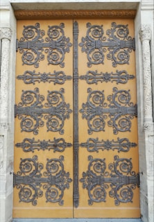 An intricately decorated wooden door featuring ornate wrought iron designs. The decorative patterns include swirling motifs and floral embellishments, creating an elaborate and detailed appearance. The door is set within a stone frame with carved columns on either side, adding to the overall grandeur.