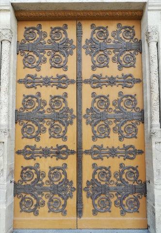 An intricately decorated wooden door featuring ornate wrought iron designs. The decorative patterns include swirling motifs and floral embellishments, creating an elaborate and detailed appearance. The door is set within a stone frame with carved columns on either side, adding to the overall grandeur.