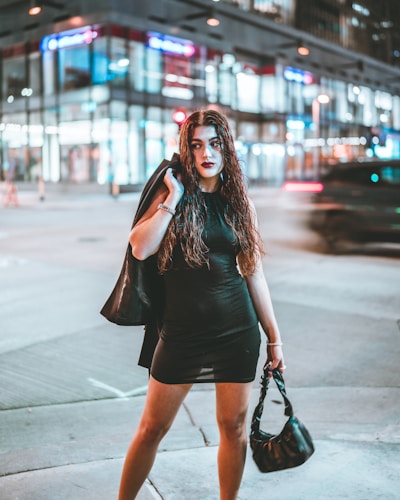 A sleek model wearing a chic, minimalist black dress standing against a cityscape backdrop at dusk.