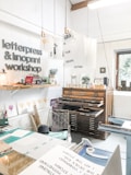 A creative studio space featuring a letterpress and linoprint workshop with printed artworks and tools. Posters with inspirational messages are suspended from the ceiling. A well-organized, artistic workspace with a wooden type cabinet and various paper materials. The room is bright and airy, with a window providing natural light.