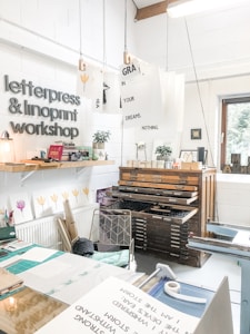 A creative studio space featuring a letterpress and linoprint workshop with printed artworks and tools. Posters with inspirational messages are suspended from the ceiling. A well-organized, artistic workspace with a wooden type cabinet and various paper materials. The room is bright and airy, with a window providing natural light.