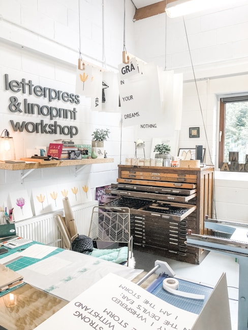 A creative studio space featuring a letterpress and linoprint workshop with printed artworks and tools. Posters with inspirational messages are suspended from the ceiling. A well-organized, artistic workspace with a wooden type cabinet and various paper materials. The room is bright and airy, with a window providing natural light.
