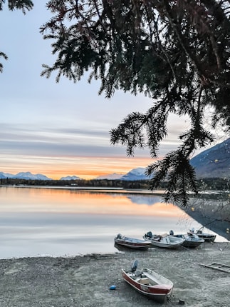 A picturesque lakeside scene at sunset, with several small boats resting on the shore. The calm water reflects the vibrant orange, pink, and blue hues of the evening sky. Pine tree branches frame the upper part of the image, and distant mountains are visible across the lake.