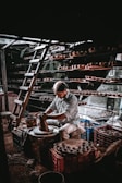 A person is working intently on a pottery wheel, shaping clay with their hands. Shelves filled with clay pots surround the workspace, and a ladder is leaning against the wall, indicating a multi-level storage setup. The lighting creates a dramatic atmosphere with shadows and highlights.