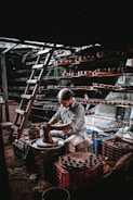 A person is working intently on a pottery wheel, shaping clay with their hands. Shelves filled with clay pots surround the workspace, and a ladder is leaning against the wall, indicating a multi-level storage setup. The lighting creates a dramatic atmosphere with shadows and highlights.
