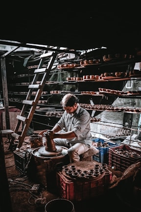 A person is working intently on a pottery wheel, shaping clay with their hands. Shelves filled with clay pots surround the workspace, and a ladder is leaning against the wall, indicating a multi-level storage setup. The lighting creates a dramatic atmosphere with shadows and highlights.