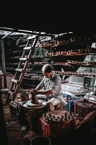 Artisan shaping clay on a pottery wheel in a sunlit studio filled with plants