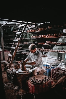 A person is working intently on a pottery wheel, shaping clay with their hands. Shelves filled with clay pots surround the workspace, and a ladder is leaning against the wall, indicating a multi-level storage setup. The lighting creates a dramatic atmosphere with shadows and highlights.