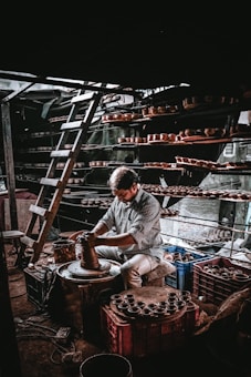 A person is working intently on a pottery wheel, shaping clay with their hands. Shelves filled with clay pots surround the workspace, and a ladder is leaning against the wall, indicating a multi-level storage setup. The lighting creates a dramatic atmosphere with shadows and highlights.