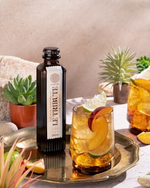 A glass of iced beverage garnished with slices of lime and peach sits on a brass tray alongside a dark bottle labeled 'Le Tribute'. The scene is complemented by small potted succulents and a sunlit background of stone and plants, creating a refreshing and natural ambiance.