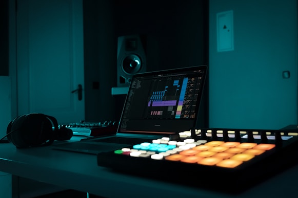 A dimly lit music production studio featuring a laptop with a digital audio workspace interface on the screen. Various music production equipment such as a MIDI controller with brightly lit pads, headphones, and a speaker is visible. The room is bathed in a cool blue light, creating a relaxed and focused atmosphere.