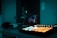 A dimly lit music production studio featuring a laptop with a digital audio workspace interface on the screen. Various music production equipment such as a MIDI controller with brightly lit pads, headphones, and a speaker is visible. The room is bathed in a cool blue light, creating a relaxed and focused atmosphere.