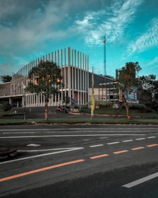A modern building with vertical strip accents on its facade, surrounded by greenery including trees and grass. A billboard with the word 'FREE' is visible nearby. There is a road in the foreground with marked lanes, and the sky is a mix of blue and cloudy patches.
