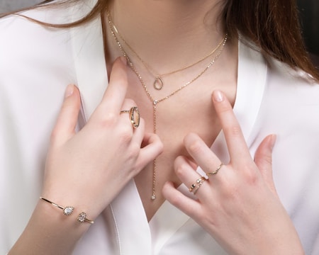 A close-up of a woman's torso showcasing delicate jewelry, including multiple gold necklaces with pendants, and several gold and gemstone rings on her fingers. She is wearing a white blouse, adding contrast to the jewelry.
