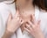 A close-up of a woman's torso showcasing delicate jewelry, including multiple gold necklaces with pendants, and several gold and gemstone rings on her fingers. She is wearing a white blouse, adding contrast to the jewelry.