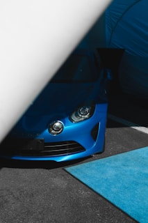A blue sports car is partially covered by a large white object, likely a tent or curtain, revealing only the front part with its headlights visible. The area has a modern, sleek aesthetic enhanced by a blue floor mat and a dark setting.