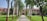 A pathway lined with palm trees leads to a red brick building with a sign that reads 'ALTERNATIVE' and other text which seems to be obscured. The courtyard is surrounded by lush green lawns and various other trees.