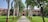 A pathway lined with palm trees leads to a red brick building with a sign that reads 'ALTERNATIVE' and other text which seems to be obscured. The courtyard is surrounded by lush green lawns and various other trees.