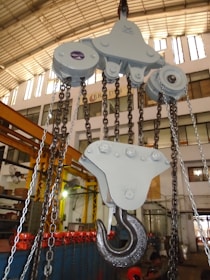 A large industrial hook attached to a pulley system is prominently displayed in a warehouse setting. The surroundings feature multiple chains and industrial equipment with a high ceiling and bright lighting.