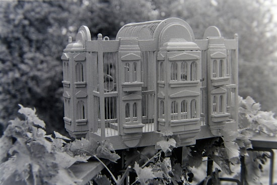 A wooden birdcage designed to resemble an elaborate building or castle, complete with detailed windows and architectural features. It is placed outdoors among lush leaves and vegetation, providing a scenic natural backdrop.