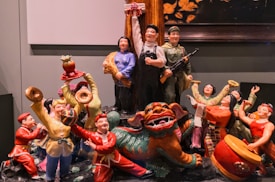 A colorful ceramic or porcelain sculpture depicting a lively scene with multiple figures engaged in joyful activities. The figures, dressed in traditional clothing, appear to be celebrating with musical instruments, a lion dance, and various expressive poses. One figure holds a large ball, while another is playing a drum. The central figure seems to hold a rifle, indicating a mix of cultural and historical themes. A dragon-like creature is prominently featured, adding to the festive mood.