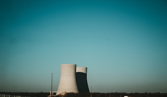 A pair of large industrial cooling towers rise against a clear blue sky, casting shadows on the ground. The towers are positioned near the horizon, with some trees and industrial structures visible at the base, along with a lamppost and fencing in the foreground.