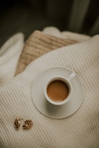 A small cup of coffee with a rich brown color sits on a white saucer placed on a cozy knitted cream-colored blanket. Next to the cup, a pair of elegant gold hoop earrings are displayed. The background also features a light brown knitted texture, creating a warm and inviting atmosphere.