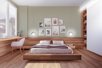 A modern bedroom with a minimalist design features a wooden platform bed with white and beige bedding. The room includes a built-in wooden desk with a white chair, a laptop, and a potted plant. A series of framed pictures are arranged on the light green wall above the bed. Pendant lights hang from the ceiling, and a bookshelf is integrated into the wall on the right, filled with books. Large windows allow natural light to illuminate the space.