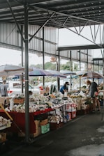 A covered market area with several stalls displaying a variety of fresh produce including vegetables and fruits. There are colorful umbrellas shading the stalls, and people are seen browsing and buying items. The market is open-air but sheltered with a metal roof structure.
