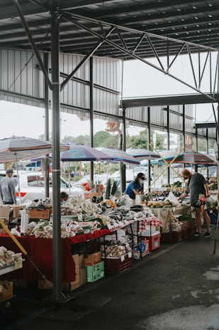 A covered market area with several stalls displaying a variety of fresh produce including vegetables and fruits. There are colorful umbrellas shading the stalls, and people are seen browsing and buying items. The market is open-air but sheltered with a metal roof structure.
