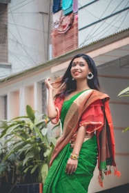 A person dressed in a traditional green and red saree stands outdoors, surrounded by lush green plants. The individual is smiling, wearing large hoop earrings, and colorful bangles. A building with hanging clothes is visible in the background.