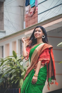 A person dressed in a traditional green and red saree stands outdoors, surrounded by lush green plants. The individual is smiling, wearing large hoop earrings, and colorful bangles. A building with hanging clothes is visible in the background.