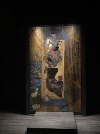 A striking artwork featuring a figure with an abstract form, surrounded by rich, colorful brushstrokes depicting a natural scene with water, lilies, and vertical elements like reeds or bamboo. The composition is set against a dark background, highlighting the vividness of the painting.