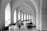A person with a cane walks down a corridor with arches, while several people lie on the floor, resting against the walls. The setting appears to be serene yet somber, with a play of light and shadows.