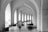 A person with a cane walks down a corridor with arches, while several people lie on the floor, resting against the walls. The setting appears to be serene yet somber, with a play of light and shadows.