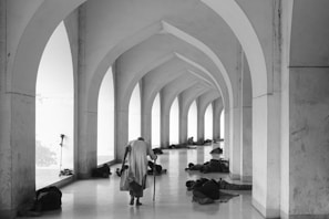A person with a cane walks down a corridor with arches, while several people lie on the floor, resting against the walls. The setting appears to be serene yet somber, with a play of light and shadows.