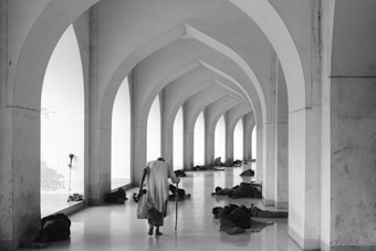 A person with a cane walks down a corridor with arches, while several people lie on the floor, resting against the walls. The setting appears to be serene yet somber, with a play of light and shadows.