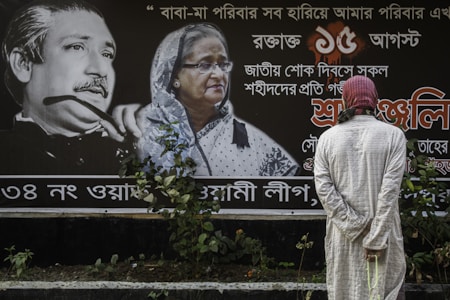 A person wearing a light-colored robe and a red and white headscarf stands in front of a large poster featuring two prominently displayed portraits accompanied by text in a script that could be Bengali. The portrait on the left shows an older man with distinctive facial features, while the portrait on the right shows an older woman wearing glasses and a white headscarf. The background of the poster is predominantly dark, with white text and accents in red.